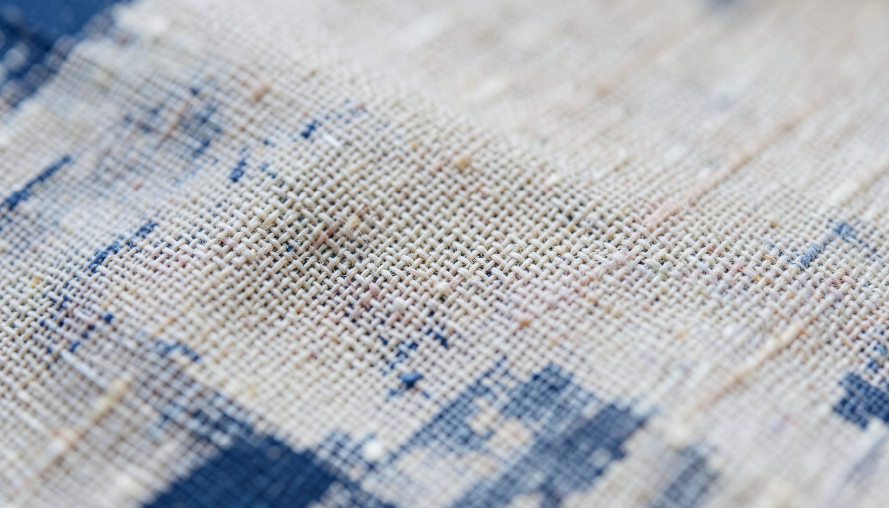 An extreme close-up photograph of woven linen fabric in natural ecru and cream tones, with a soft diffuse light revealing the fine thread texture and subtle variations in the warp and weft.
